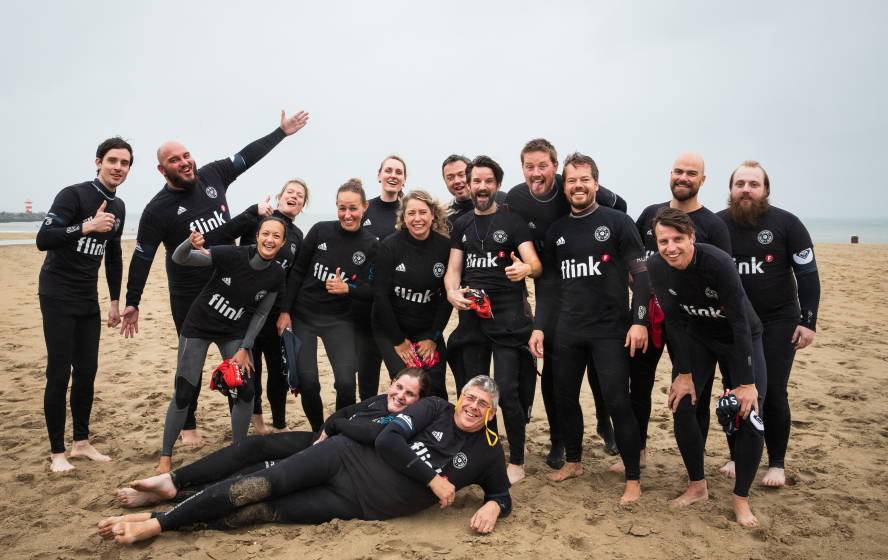 Teamuitje groepsfoto van alle Flinkerts in wetsuits op het strand